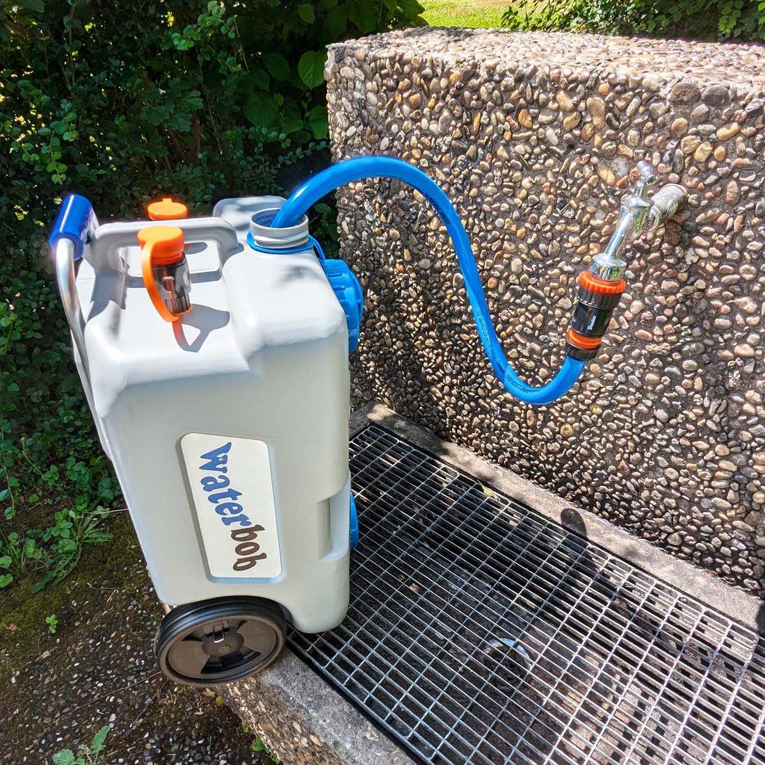 WaterBob steht an einem Wasserhahn und wird über den blauen Schlauch gefüllt